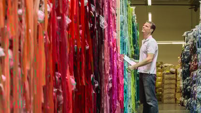 Man inspecting rolls of colorful fabric in a warehouse, holding a clipboard